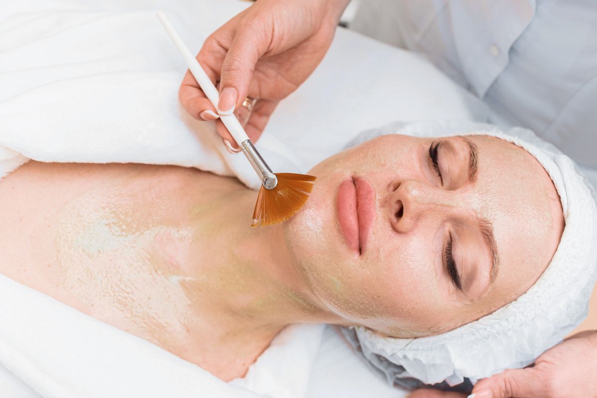 A imagem mostra uma mulher de olhos fechados num momento de tratamento estético facial, como peeling químico, enquanto um profissional aplica uma máscara facial com um pincel macio. A cena, em um ambiente de spa ou clínica, transmite uma sensação de cuidado profissional e tranquilidade.