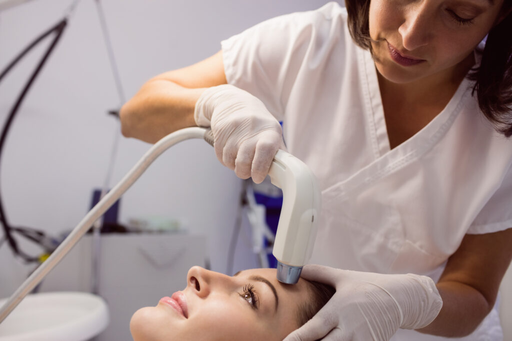Profissional de saúde realiza procedimento estético facial em uma mulher deitada, utilizando um aparelho aplicado na região da testa.