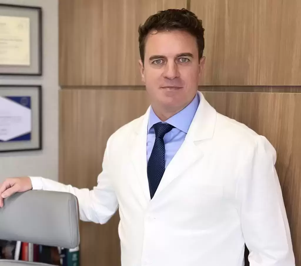 Retrato profissional do dermatologista Paulo Junqueira em consultório, vestindo jaleco branco e gravata azul, com diplomas na parede ao fundo.