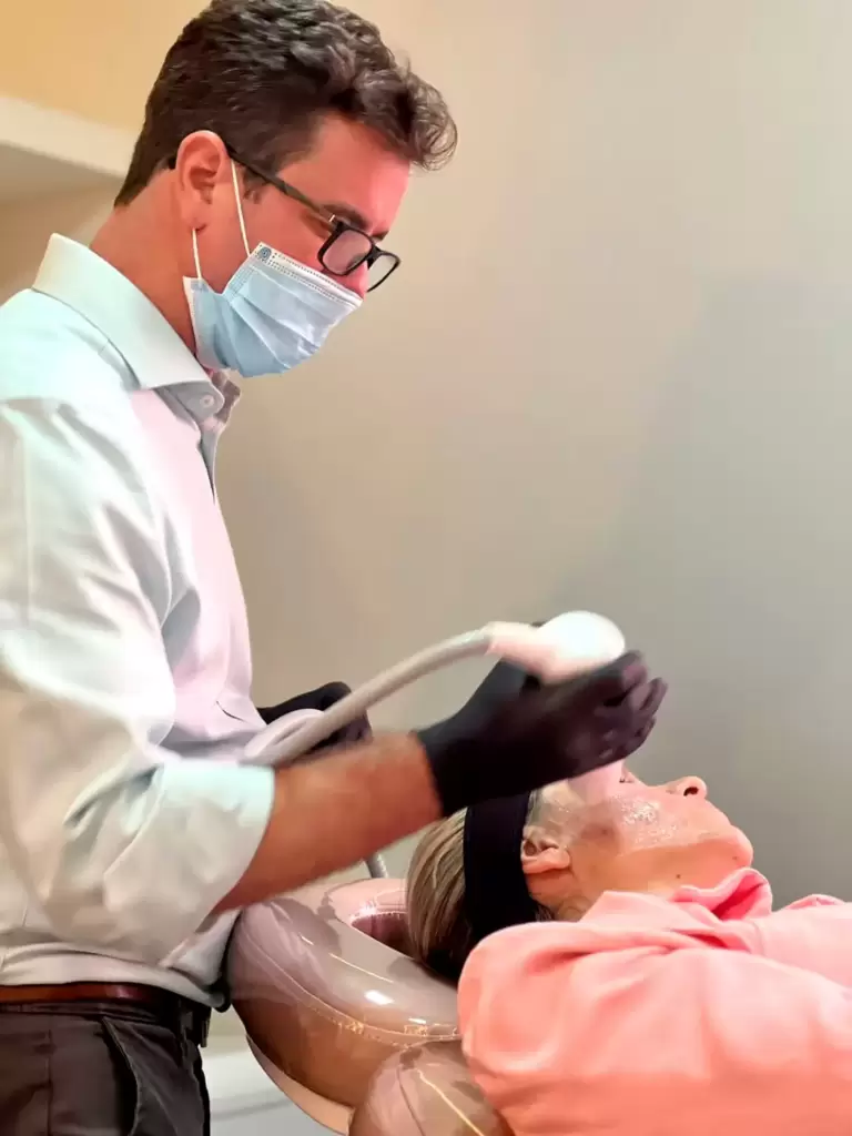 Dermatologista Dr. Paulo Junqueira realiza procedimento facial com aparelho dermatológico em paciente deitada em clínica.